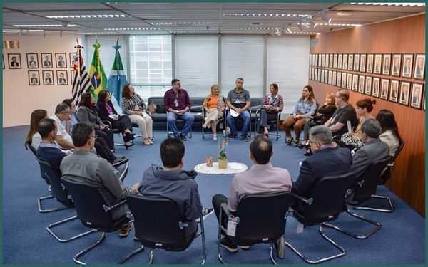 A imagem mostra um grupo de cerca de doze pessoas sentadas em círculo em uma sala, em cadeiras organizadas de forma que todos possam se ver, sugerindo uma reunião ou atividade em grupo. No centro, há um pequeno tapete claro com um apoio contendo objetos decorativos, como plantas. Algumas pessoas seguram papéis ou cadernos. O ambiente é bem iluminado por uma janela com persianas ao fundo, e há várias molduras alinhadas na parede. O chão é coberto por carpete azul, e a cena transmite um espaço organizado voltado para conversa ou dinâmica coletiva.